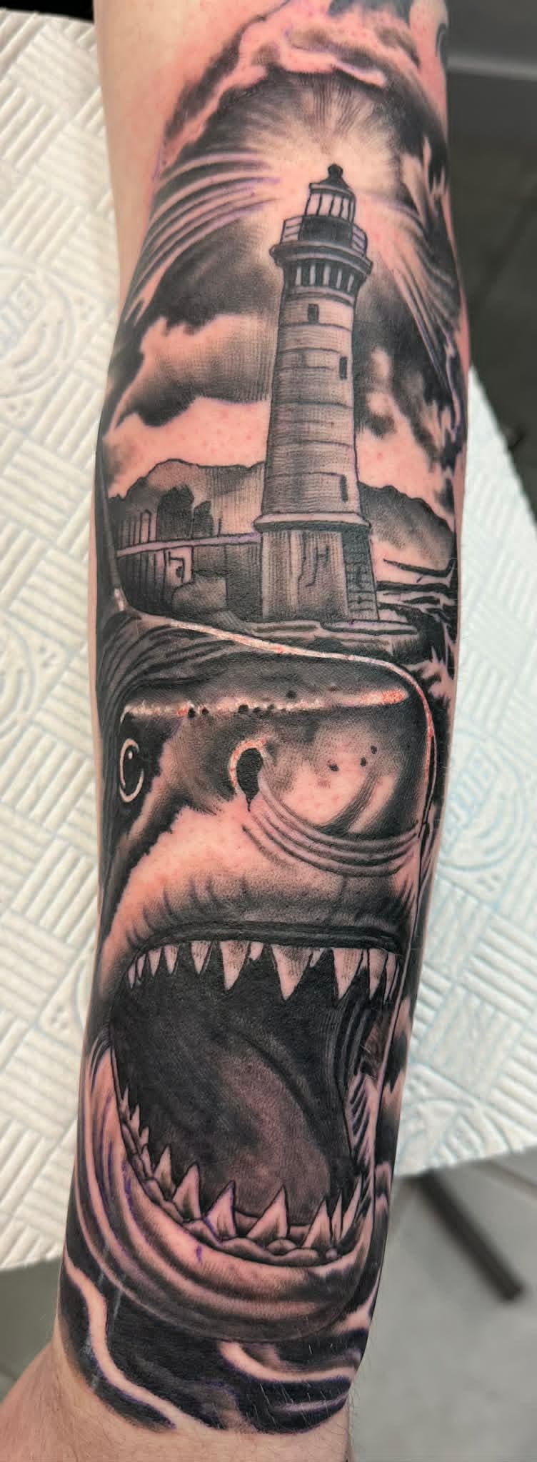 Realism lighthouse on rocky shore with great white shark breaching below forearm tattoo by John Quinn at Tattoo Zone Newcastle.