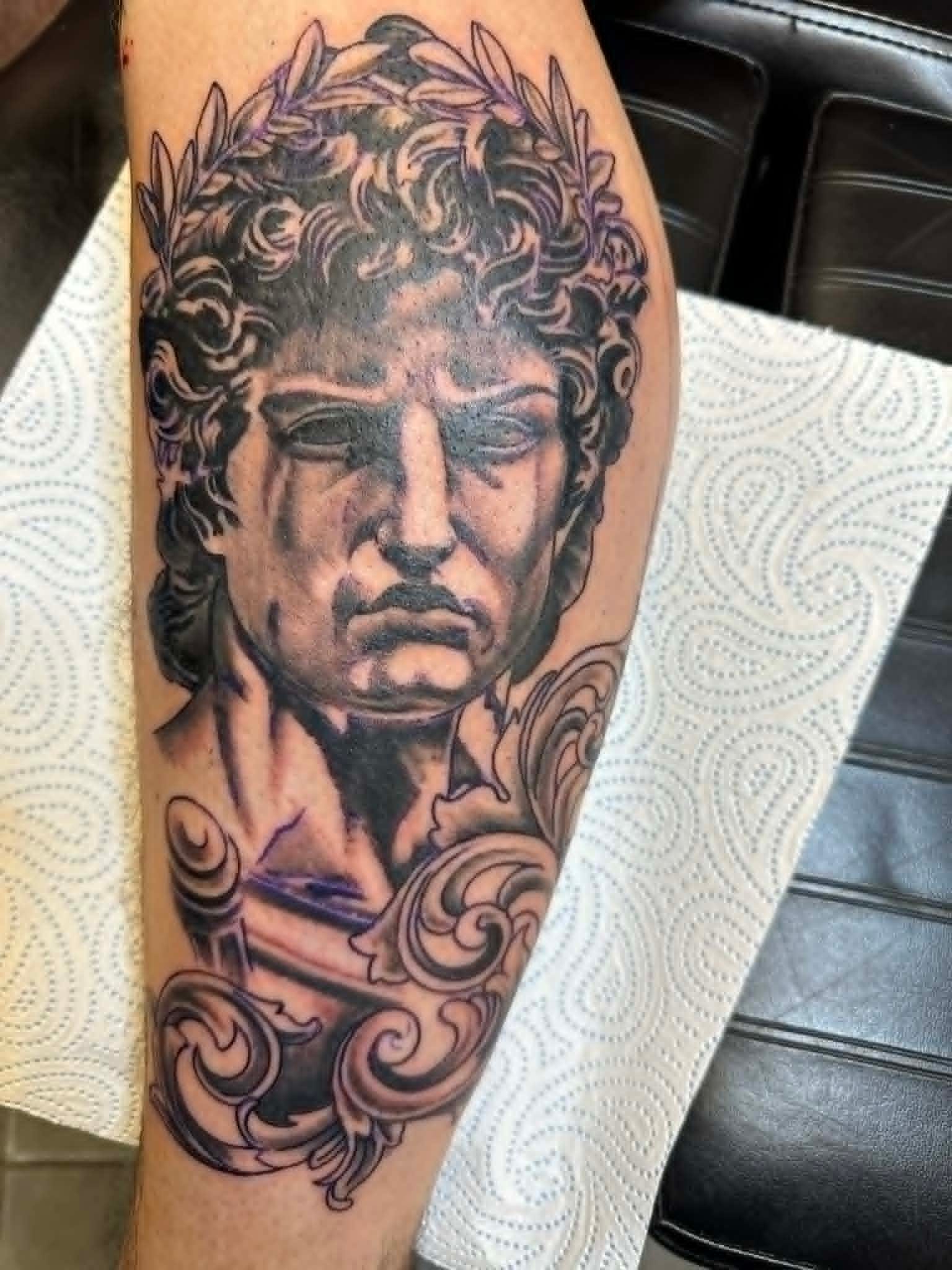 Black and grey Greek statue portrait with laurel wreath and filigree tattoo on the forearm by John Quinn at Tattoo Zone Newcastle.